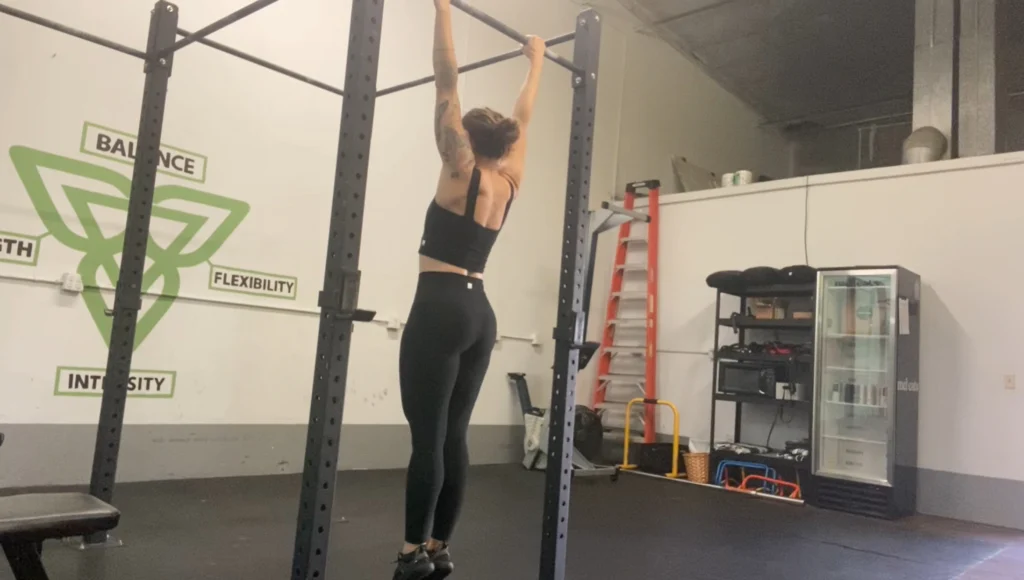 A woman demonstrating how to perform the seated scapula pull-up on a chin-up bar.